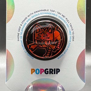 May include: A black phone grip with a red and black cartoon character design. The grip has a swappable top and says "Try Me! Pull Twice to Grip" and "Essayer-moi! Tirez deux fois pour saisir" in English and French. The grip also says "PopGrip" on the bottom.
