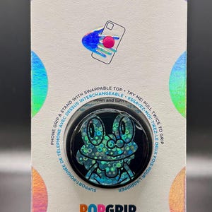 May include: A PopSockets PopGrip featuring a black base with a frog design in shades of blue and green. The packaging includes the PopSockets logo and text: "PHONE GRIP & STAND WITH SWAPPABLE TOP".