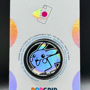 Può includere: Un PopSockets PopGrip con un design olografico di Pikachu. L'accessorio per telefono ha un bordo nero ed è attaccato a un supporto bianco con i loghi PopSockets e PopGrip. La confezione include testo in inglese e francese.