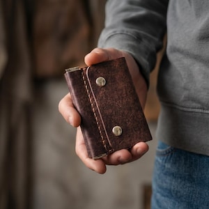 Op de afbeelding: Een bruine leren sleutelhouder met een getextureerde afwerking, vastgezet met twee goudkleurige druksluitingen. De sleutelhouder wordt in een hand gehouden, wat het compacte ontwerp en vakmanschap laat zien.