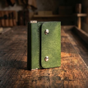 Pode incluir: Um porta-chaves de couro verde com fecho de aba preso por dois rebites prateados. O porta-chaves é feito de couro texturizado e tem uma borda costurada. É exibido em uma superfície de madeira.