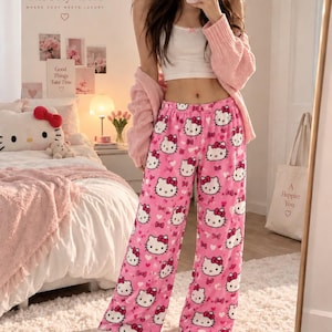 May include: Pink plush pyjama trousers featuring an all-over pattern of Hello Kitty faces and small hearts. The trousers have a wide-leg silhouette and an elastic waistband. The fabric appears to be a soft, fuzzy material suitable for loungewear.