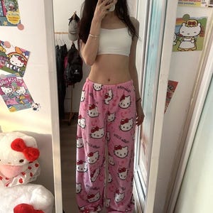 Puede incluir: Pantalones de pijama rosas con un estampado repetido de caras y lazos de Hello Kitty. Los pantalones se combinan con zapatillas Hello Kitty a juego y un top corto blanco. La imagen est&aacute; tomada frente a un espejo.