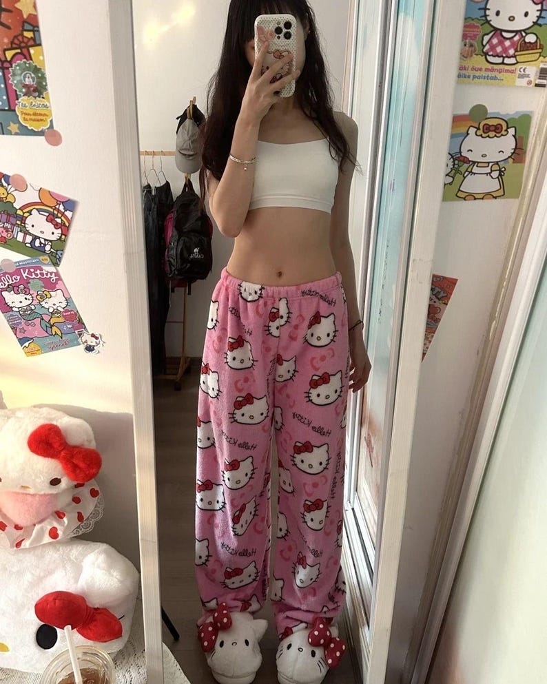 Puede incluir: Pantalones de pijama rosas con un estampado repetido de caras y lazos de Hello Kitty. Los pantalones se combinan con zapatillas Hello Kitty a juego y un top corto blanco. La imagen est&aacute; tomada frente a un espejo.
