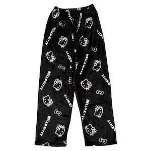 Puede incluir: Pantalones de pijama negros con un patr&oacute;n repetido de caras de Hello Kitty blancas, lazos y las palabras "Hello Kitty". Los pantalones est&aacute;n hechos de un material suave y afelpado.