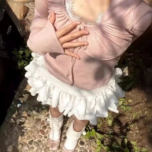 May include: A light pink long-sleeved top with a lace trim neckline and a white ruffled skirt. The outfit includes matching pink shoes with bows and white lace socks. The clothing is suitable for a casual or dressy occasion.