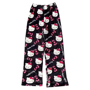 Puede incluir: Pantalones de pijama negros con un patr&oacute;n repetido de caras de Hello Kitty, lazos rosas y las palabras "Hello Kitty" en blanco. Los pantalones est&aacute;n hechos de un material suave y afelpado.