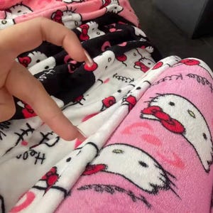 Puede incluir: Primer plano de un pantal&oacute;n de pijama de felpa rosa, negro y blanco. La tela presenta un patr&oacute;n repetido de un gato de dibujos animados con un lazo rojo. Las palabras "Hello Kitty" est&aacute;n impresas en la tela. El pijama parece suave y esponjoso.