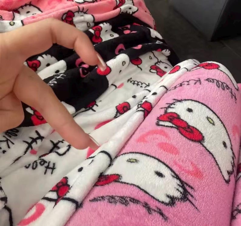 Puede incluir: Primer plano de un pantal&oacute;n de pijama de felpa rosa, negro y blanco. La tela presenta un patr&oacute;n repetido de un gato de dibujos animados con un lazo rojo. Las palabras "Hello Kitty" est&aacute;n impresas en la tela. El pijama parece suave y esponjoso.