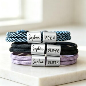 Puede incluir: Tres pulseras de cordón trenzado en azul y negro, negro sólido y morado claro. Cada pulsera cuenta con dos cuentas rectangulares de metal plateado grabadas con los nombres Sophia, Oliver o el año 2024 en letras negras.
