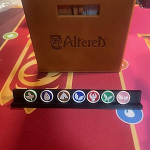 May include: A brown leatherette card storage box with the word "Altered" printed on the front. A black plastic card holder with seven circular tokens with various symbols is in front of the box. The tokens are green, blue, and pink.