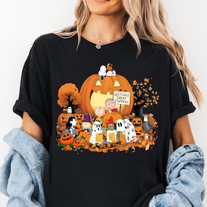 May include: A black short-sleeve t-shirt featuring a colorful graphic of Peanuts characters dressed in Halloween costumes. The design includes a large orange pumpkin, smaller pumpkins, autumn trees, and a sign that reads Welcome Great Pumpkin.