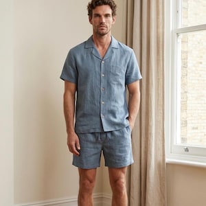 Peut inclure: Un ensemble de pyjama en lin bleu clair, composé d'une chemise à manches courtes boutonnée et d'un short assorti. La chemise a une poche de poitrine. L'ensemble est présenté dans une pièce lumineuse avec une fenêtre et des rideaux beiges.