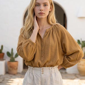 Pode incluir: Uma blusa de manga comprida amarelo-mostarda com carcela de botões e decote franzido, combinada com calças bege-claras de cintura subida com cós tipo paperbag e três botões visíveis na parte central frontal.