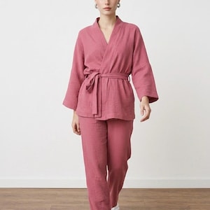 May include: A dusty rose two-piece lounge set featuring a long-sleeved wrap-style top with a matching tie belt and coordinating straight-leg trousers. The garment is crafted from a textured, crinkled fabric and is paired with white slide sandals.