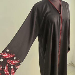 Op de afbeelding: Een donkerbruine abaya met rode bies langs de halslijn en de voorkant. De mouwen hebben een geborduurd ontwerp van rode en witte veren en kleine witte bloemen. De abaya wordt getoond op een mannequin.