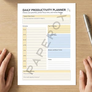 May include: A white Daily Productivity Planner sheet with a schedule, priority list, and notes section. The planner includes the text "Choose your priorities, protect focus time, and review the day." A silver pen rests on the wooden surface.