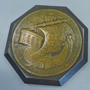 Puede incluir: Una medalla de bronce octogonal con un barco con un edificio en su cubierta, una corona y el texto "TIENDE NEDERLANDSCHE JAARBLIJDSU PRECHT MAART 1924".