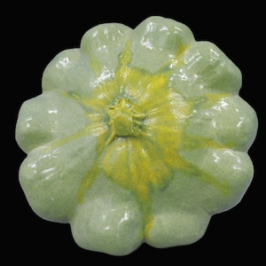 Op de afbeelding: Een keramisch decoratief object in de vorm van een pompoen. Het object is lichtgroen van kleur met een gele kern. De pompoen heeft een textuuroppervlak en staat tegen een zwarte achtergrond. De pompoen is een decoratief item.