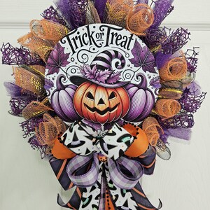 May include: A Halloween wreath with a central circular design featuring a smiling jack-o'-lantern wearing a witch's hat, flanked by purple pumpkins. The words "Trick or Treat" are above the pumpkin. The wreath is decorated with orange, purple, and black ribbons and mesh.