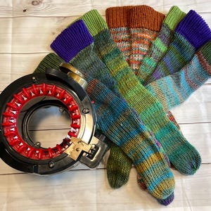 May include: A black and red circular knitting machine with several pairs of colorful, hand-knitted socks. The socks feature a variety of colors, including green, blue, purple, and orange. The socks are made of yarn and are lying on a wooden surface.