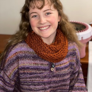 May include: A young person wearing a purple and brown striped sweater with a brown knitted scarf. The person is smiling and looking at the camera.