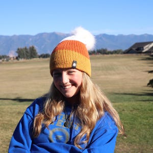 Può includere: Un berretto lavorato a maglia arancione, giallo e bianco con un soffice pompon bianco. Il cappello ha una piccola etichetta nera. La persona indossa una felpa blu. Montagne e un campo sullo sfondo.