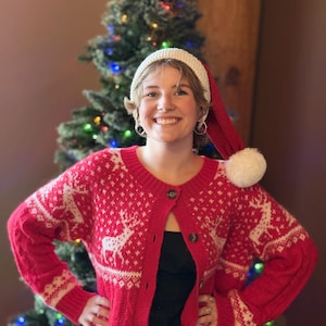 Puede incluir: Un cárdigan rojo con un estampado de renos blancos y cierre de botones. La persona lleva un gorro de Papá Noel blanco con borde rojo y pompón blanco. Un árbol de Navidad está en el fondo.