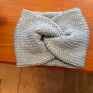 May include: A light blue knitted headband with a twisted knot design. The headband is made of a soft, textured yarn and is designed to keep the ears warm. The headband is approximately 9 inches wide.