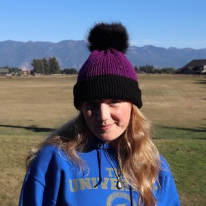 Op de afbeelding: Een gebreide beanie-muts met een zwarte basis en een paarse bovenkant, bekroond met een zwarte imitatiebont pompon. De muts wordt gedragen door een persoon met lang blond haar, tegen een achtergrond van een veld, bergen en een heldere blauwe lucht.