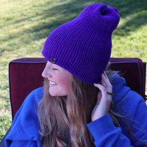 May include: A vibrant purple knit beanie with a ribbed texture. The hat has a small, dark brown tag on the side. The person in the image is wearing a blue sweatshirt.