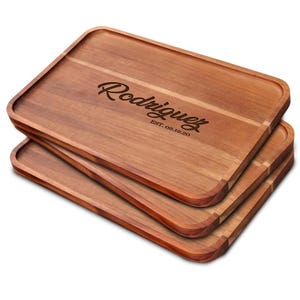 May include: Three rectangular wooden serving trays stacked on top of each other. The top tray has the name "Rodriguez" engraved in a cursive font, with "EST. 02.12.20" below it. The trays are a warm brown color.