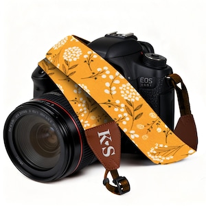 May include: A black DSLR camera with a mustard yellow camera strap featuring a white floral pattern. The strap includes brown leather-like end tabs with the white letters K and S separated by a small heart symbol, and black plastic buckles.
