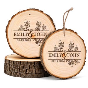 May include: Two wooden ornaments with bark edges, engraved with the names "EMILY & JOHN" and the date "02.15.2024", along with leaf designs. One ornament has a twine loop for hanging. A stack of wood slices is in the background.