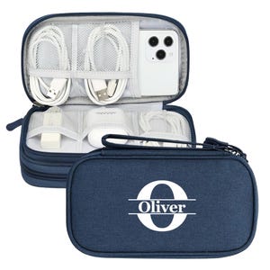 May include: Navy blue travel organizer with white cables, a phone, and accessories. The case is open, revealing compartments for electronics. The exterior features the name "Oliver" with a large initial "O".