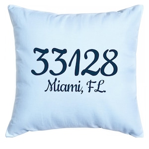 May include: A light blue square pillow with the dark blue text "33128 Miami, FL." The pillow is a solid light blue color and has a soft, textured appearance. The text is in a cursive font.