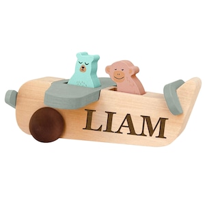 May include: Wooden toy airplane with a light brown body, gray accents, and the name "LIAM" printed on the side. It features a teal bear and a pink monkey as passengers. The propeller is brown.