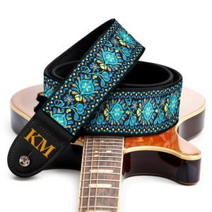 May include: A guitar strap featuring a vibrant teal, blue, and yellow floral pattern on a black background. The strap has black leather ends, one of which displays the gold letters KM and a silver metal button. It rests on a wooden guitar body.