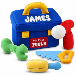 May include: A blue plush toy toolbox with a yellow handle, featuring the name JAMES in white letters and a red patch labeled My First TOOLS. Included are a green wrench, a red hammer with a white head, a blue saw, and a yellow screwdriver.