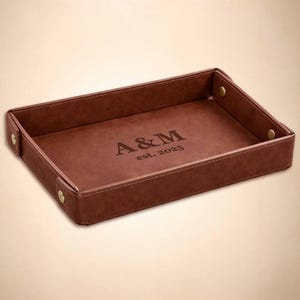May include: A rectangular brown valet tray with raised sides secured by brass-colored metal snaps at each corner. The center of the tray features the engraved text A&M and est. 2025 in a serif font, providing a structured surface for small items.
