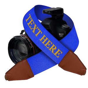 May include: A blue camera strap with brown leather ends. The strap has the text "TEXT HERE" embroidered in gold.