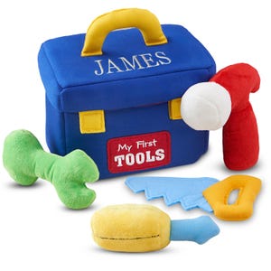 May include: A blue fabric toy toolbox with a yellow handle and the name "JAMES" embroidered on the front. The toolbox includes toy tools: a green wrench, a red hammer, a blue saw, and a yellow screwdriver. The toolbox has a red label that says "My First TOOLS".