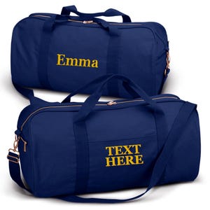 May include: Two navy blue canvas duffel bags with gold zippers and straps. The top bag has the name "Emma" embroidered in gold on the front. The bottom bag has the text "TEXT HERE" embroidered in gold on the front.