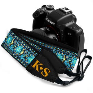 May include: Black camera strap with a blue, yellow, and teal floral pattern. The strap has the initials "K❤️S" embroidered in gold. A black Canon camera is in the background.