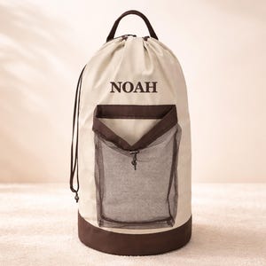 May include: A beige drawstring laundry bag with a brown base, handle, and pocket. The bag features the name "NOAH" in dark brown lettering. The pocket is made of a dark mesh material and has a drawstring closure.