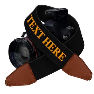 May include: Black camera strap with brown leather ends. The strap has the text "TEXT HERE" embroidered in yellow.