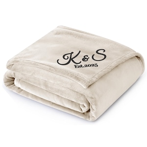 May include: Folded beige blanket with the embroidered initials "K&S" and the year "Est.2025" in black script. The blanket is made of a soft, plush material and is folded neatly, showcasing its texture and design.