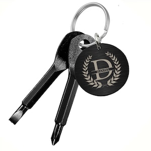 May include: Black multi-tool keychain with a flathead screwdriver, Phillips head screwdriver, and a round tag. The tag is engraved with the name "DANIEL" and the letter "D" inside a laurel wreath. The keychain is attached to a silver ring.