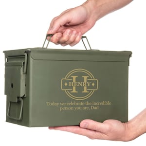 May include: An olive green metal ammo box with a handle is held by two hands. The box features a gold emblem with the name "HENRY" and the text "Today we celebrate the incredible person you are, Dad."