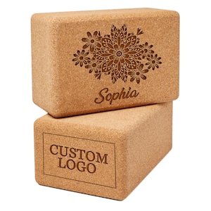 May include: Two rectangular cork yoga blocks. The top block features a floral design and the name "Sophia" engraved. The bottom block has the text "CUSTOM LOGO" engraved. The blocks are a natural tan color.